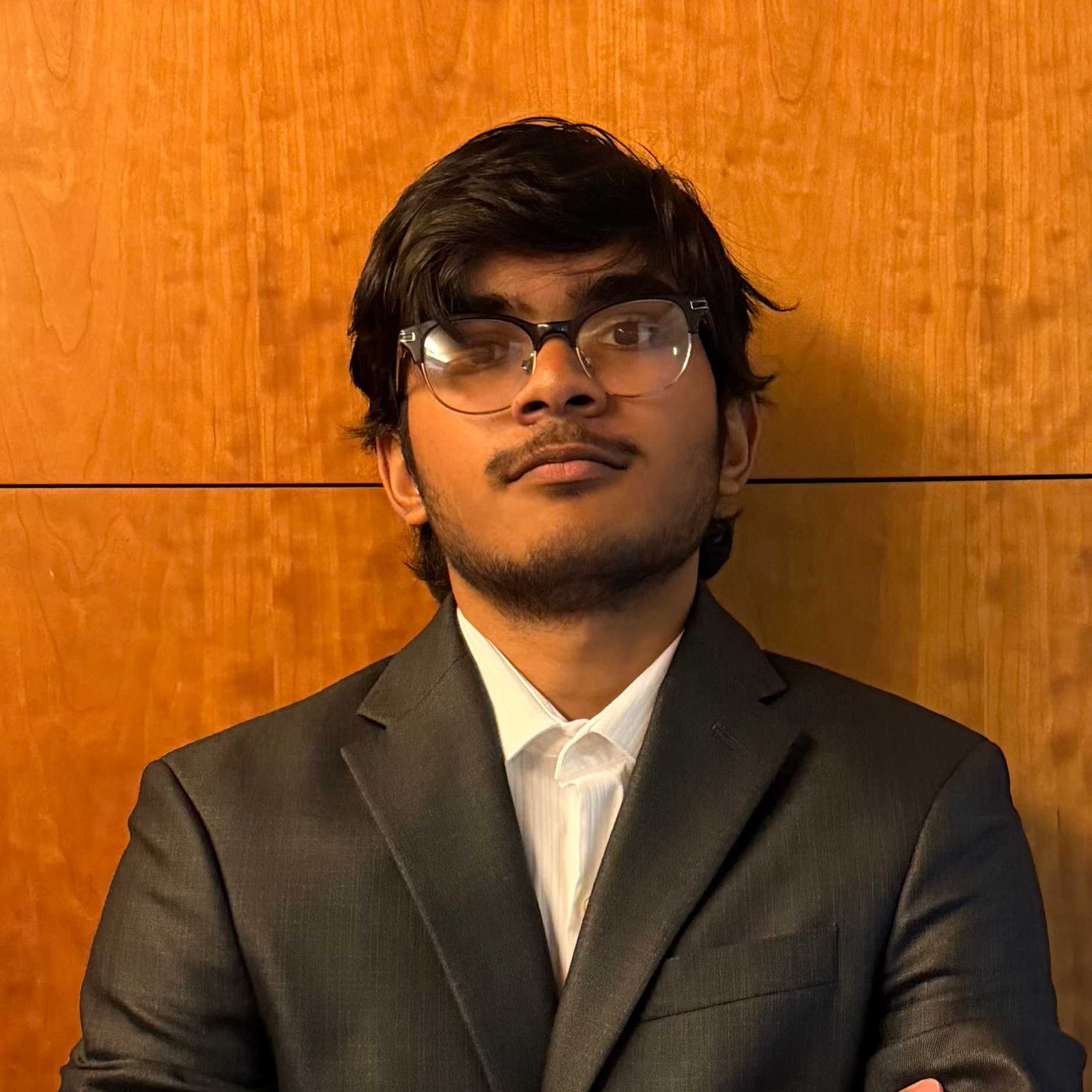 A young man wearing a suit jacket and glasses appears leaning upon a wal.