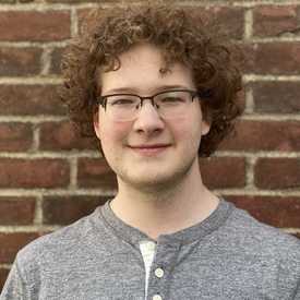 A smiling person with red curly hair appears in front of a brick wall.