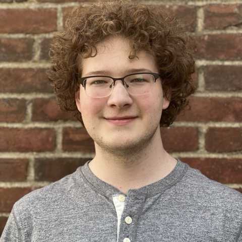 A smiling person with red curly hair appears in front of a brick wall.