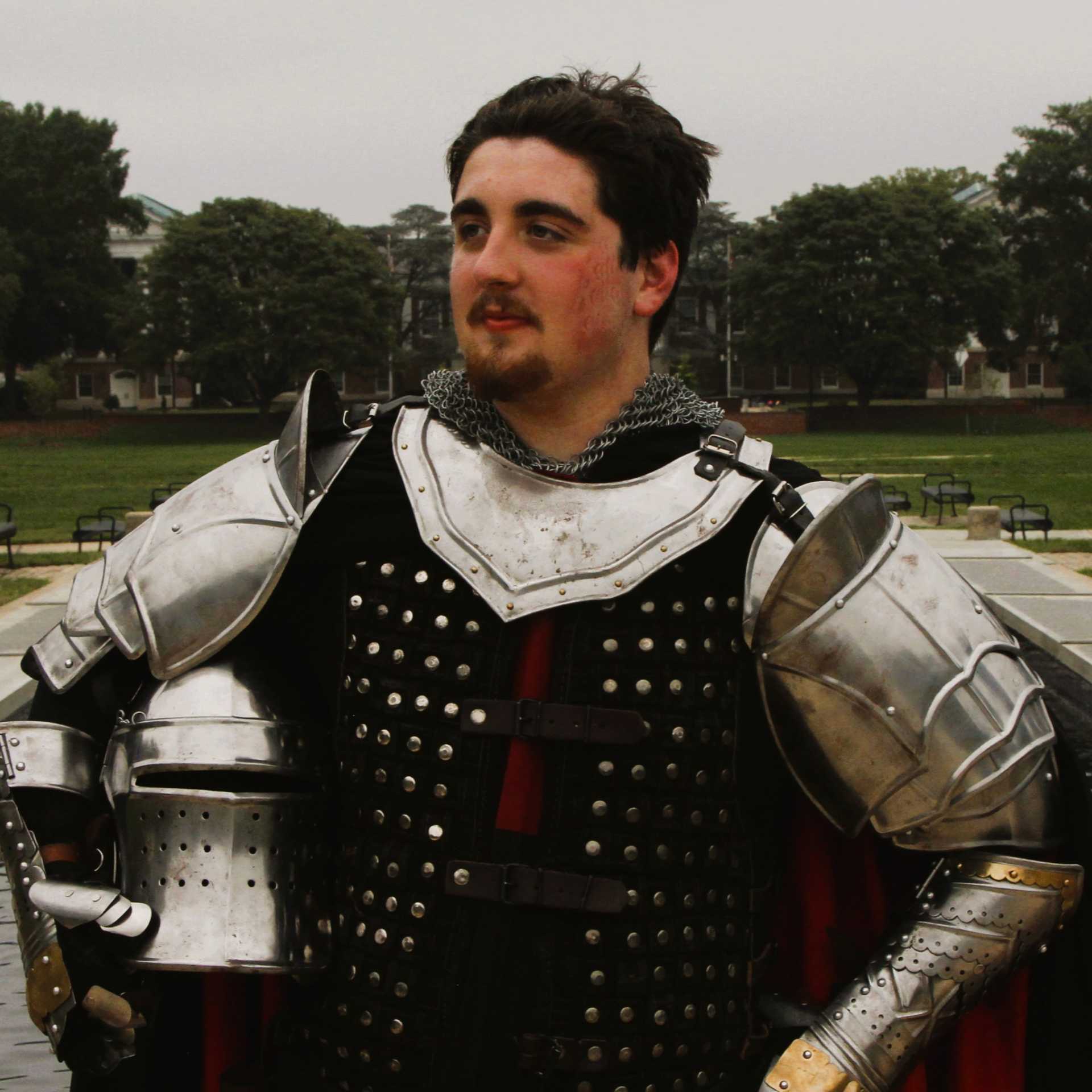 A young man in medieval armour stands proudly, holding his helmet.