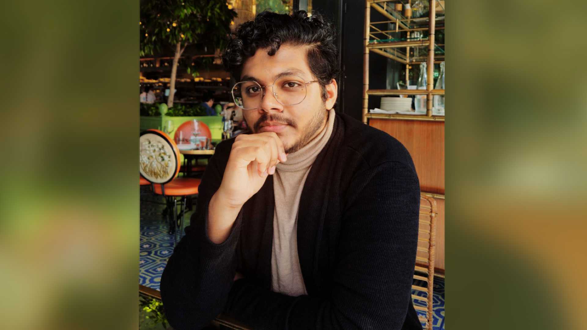 A young man with dark hair wearing a blazer and glasses appears in a restaurant setting.