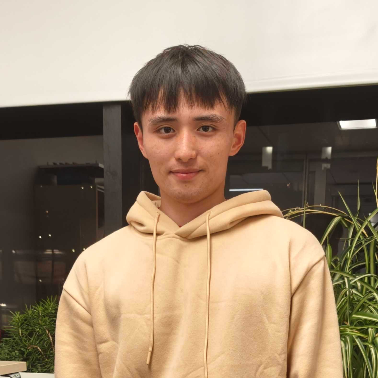 A young man appears in a yellow hoodie, with dark hair and a slight smile