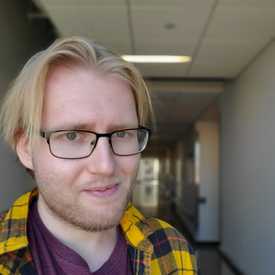 A young man with blond hair and glasses, in a hallway, smiles at the camera.