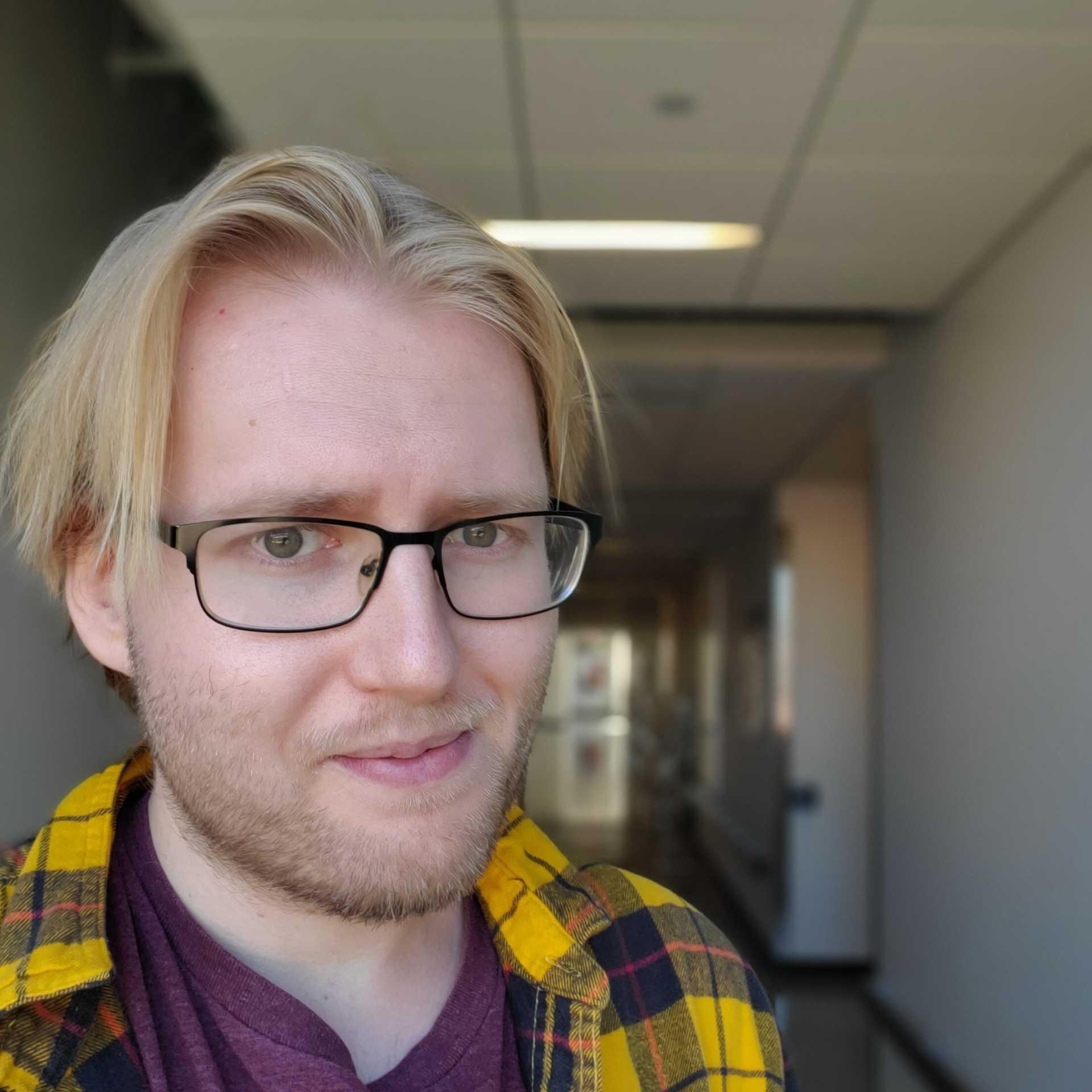 A young man with blond hair and glasses, in a hallway, smiles at the camera.