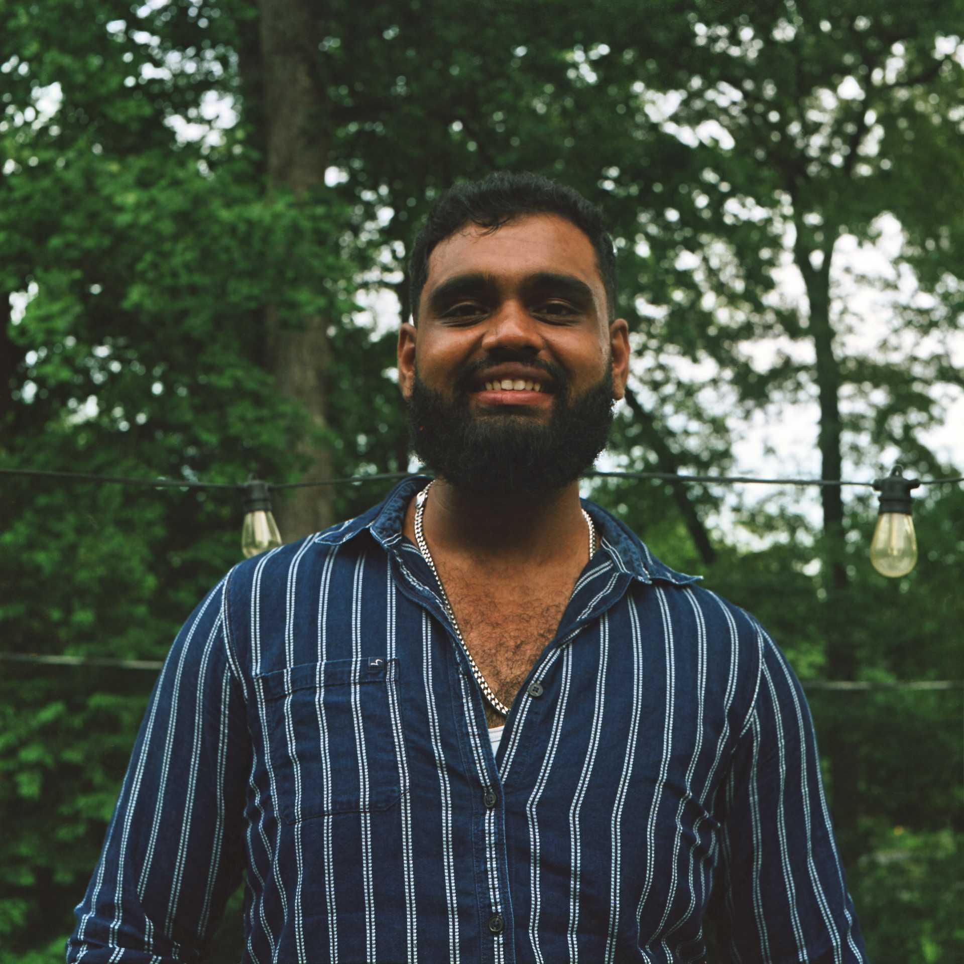 A darl-haired, bearded young man in the outdoors appears smiling; 