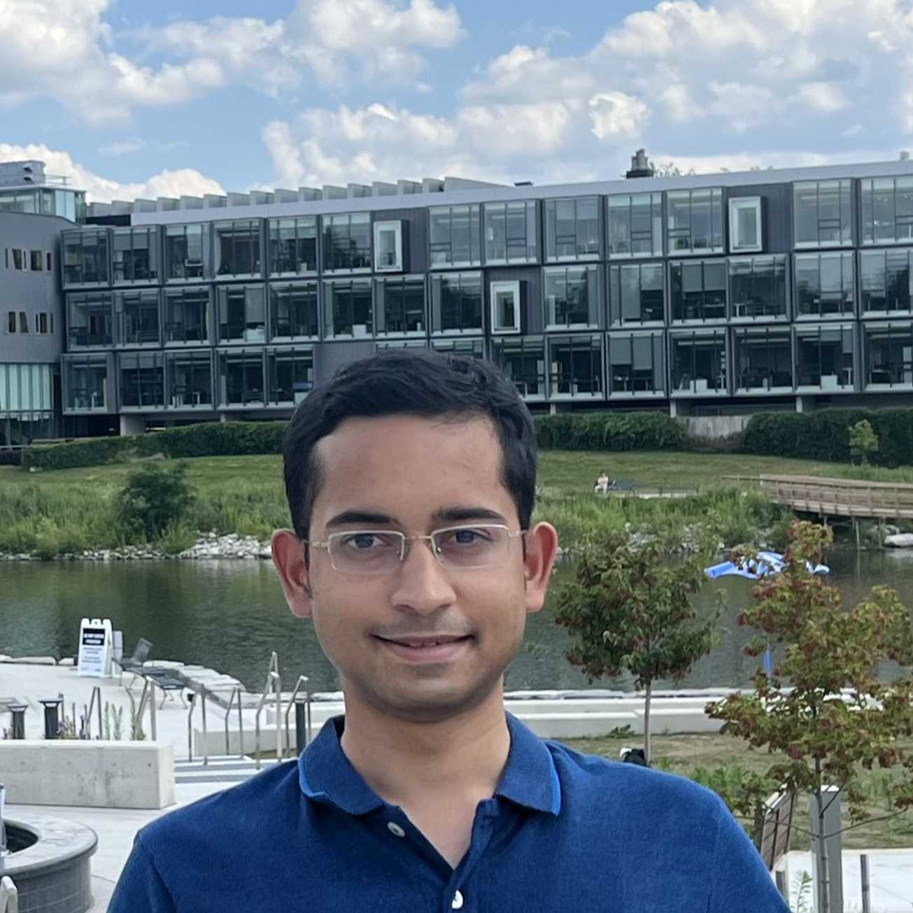 A smiling young man with brown skin and short hair is picutred in an outdoor environment.