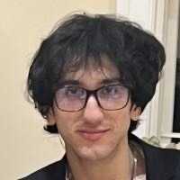 A young man wearing glasses appears indoors, smiling.